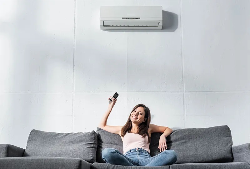 A woman sits on a couch holding the remote that controls her ductless heat pump.